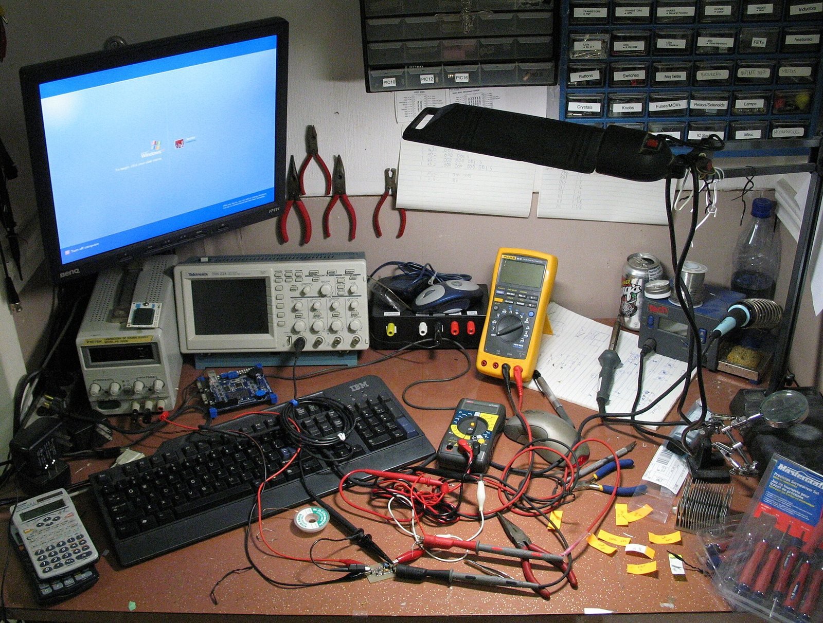 Crowded electronics workbench with oscilloscope, meters, and soldering tools